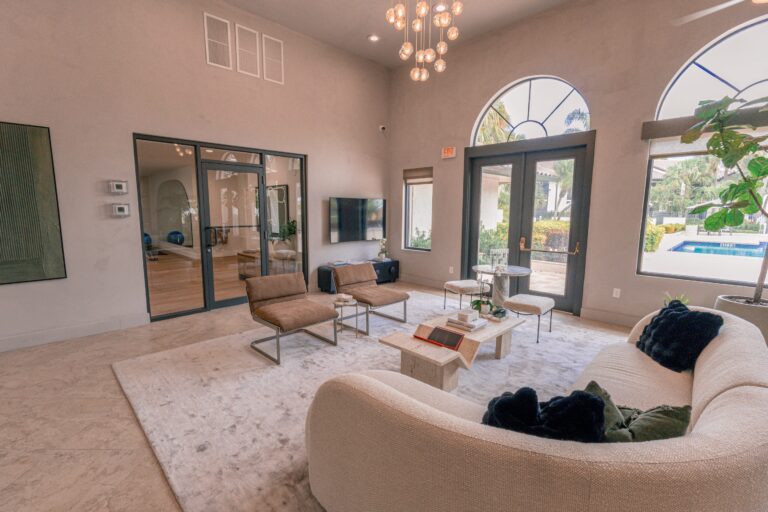 Modern living room with a curved sofa, two brown chairs, marble coffee table, wall-mounted TV, and large windows overlooking a pool and garden.