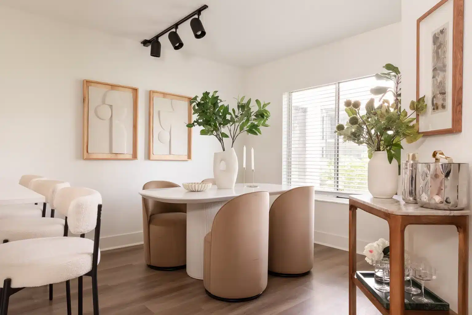 Modern dining room with a round white table, four beige chairs, abstract wall art, potted plants, and a window with blinds letting in natural light.